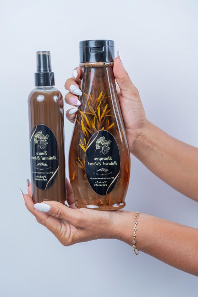 Una persona sostiene un frasco pulverizador marrón y un frasco transparente que contiene hierbas, ambos con etiquetas negras de la misma marca.