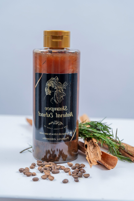Una botella de champú marrón con tapón dorado reposa sobre una superficie blanca con granos de café esparcidos, una ramita y canela.