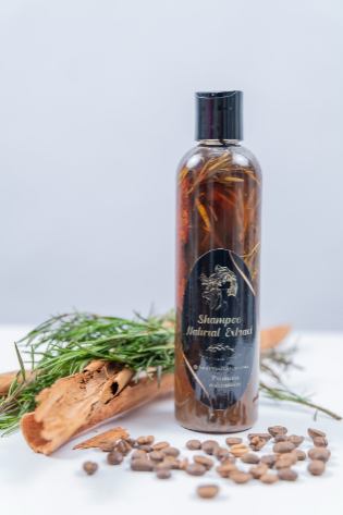 Una botella de champú con ingredientes herbales, ramas de canela y granos de café sobre una superficie blanca.