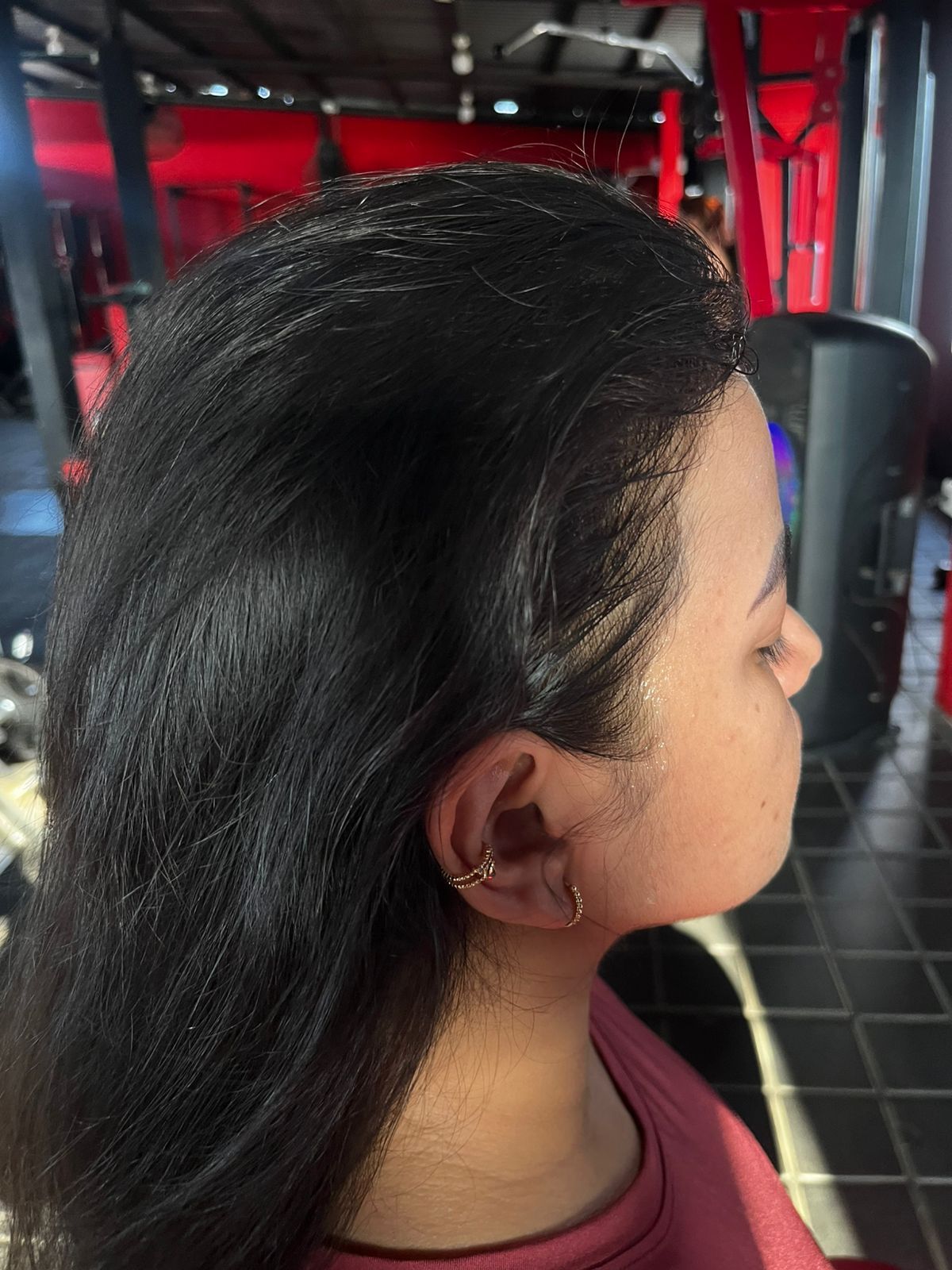 Vista de perfil de una persona con cabello oscuro, que lleva un pequeño pendiente de aro dorado, dentro de un gimnasio con detalles en rojo.