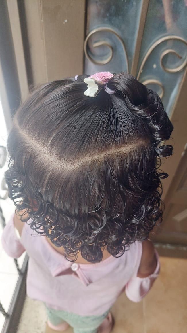 Primer plano, tomado desde un ángulo elevado, del cabello oscuro y rizado de una persona, peinado con un accesorio floral rosa y la raya al lado.