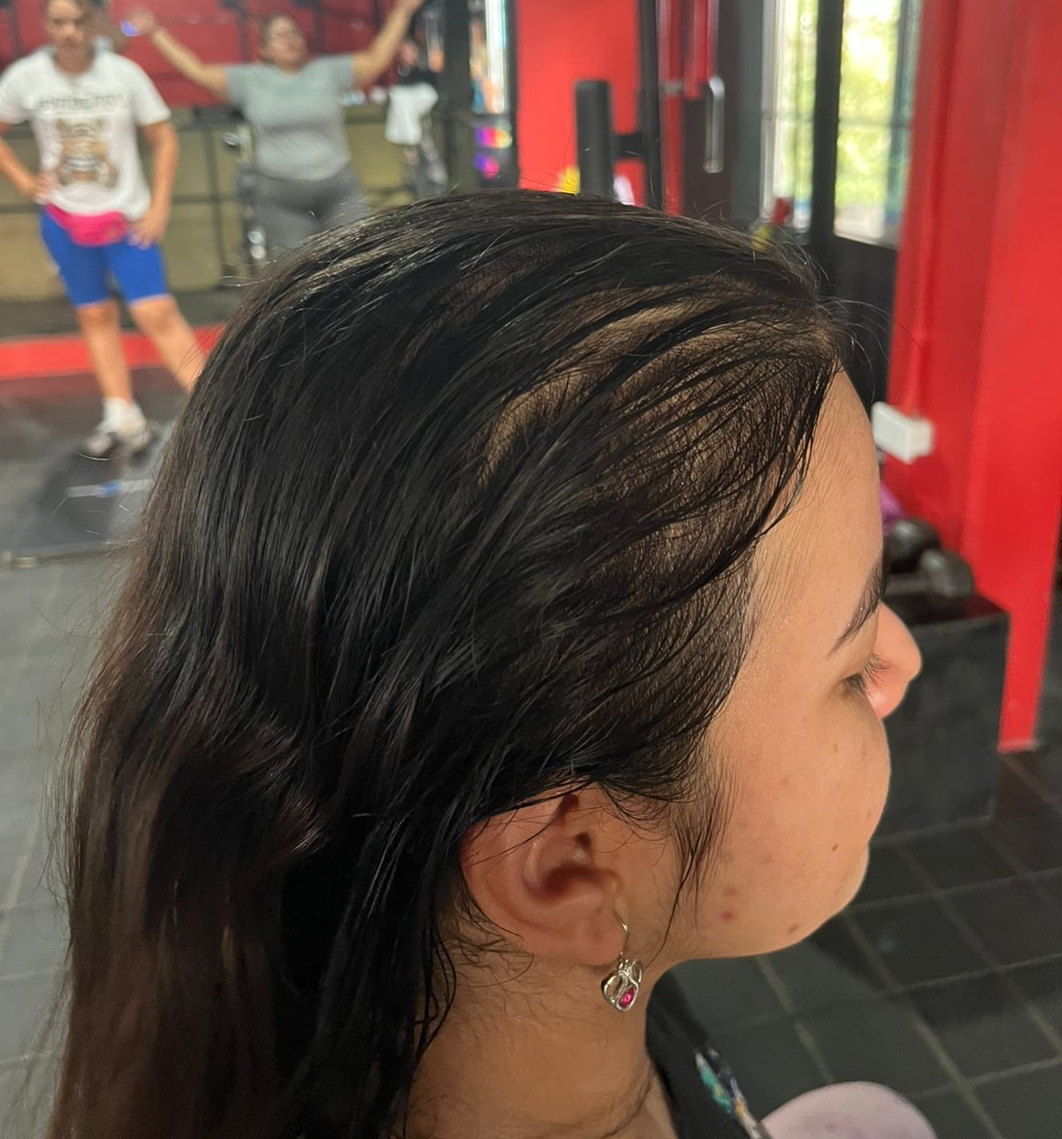 Vista lateral de una persona con cabello oscuro en un gimnasio, con otra persona haciendo ejercicio al fondo, desenfocada.