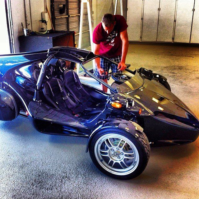 Man inspecting a dark blue three-wheeled vehicle in a garage.