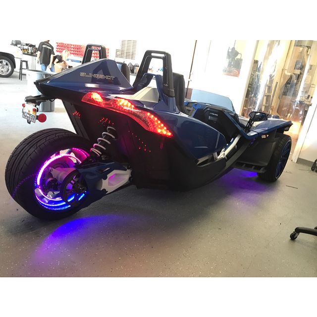 Blue three-wheeled Slingshot with glowing purple lights on a shop floor.