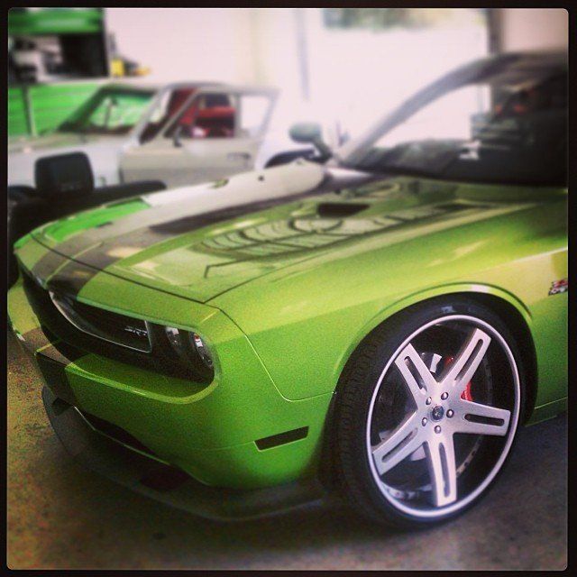 Green Dodge Challenger with black stripes and custom wheels in a garage, another car in background.