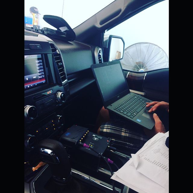 Inside a vehicle, a person uses a laptop connected to devices, papers in lap; white fan visible.