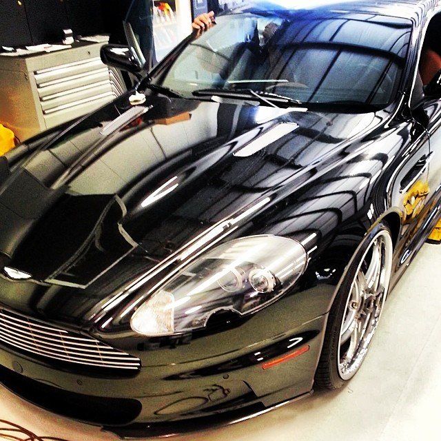 Black Aston Martin sports car in a garage, reflecting the setting; car's sleek hood and chrome wheels are highlighted.