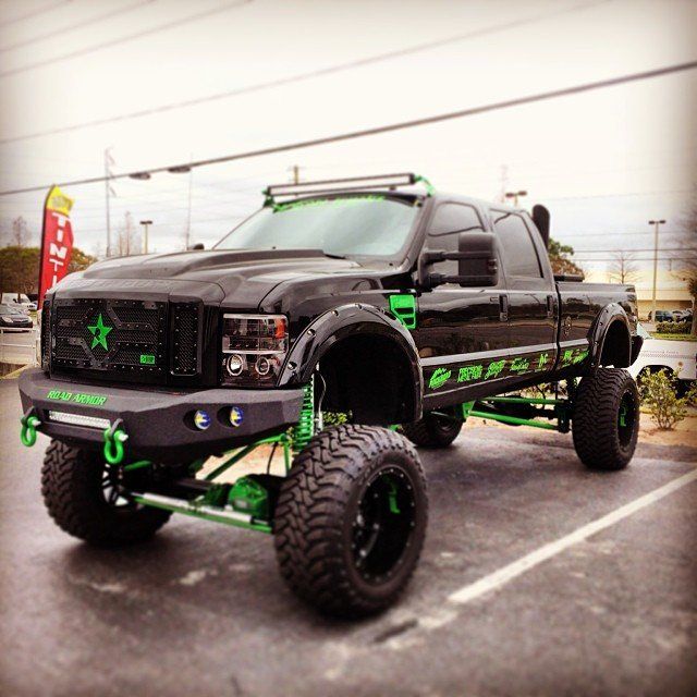 Black and green lifted truck with oversized tires parked on a paved area, with a light bar and custom grill.