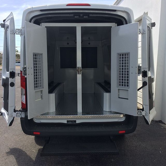 White van with open doors, interior jail cell structure visible. Rear door ramp extended.