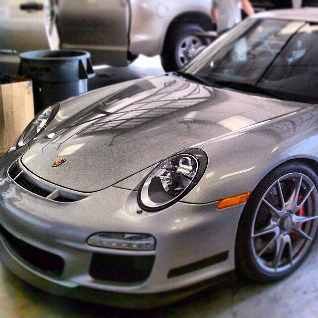 Silver Porsche sports car in a garage, headlights on, parked near a truck.