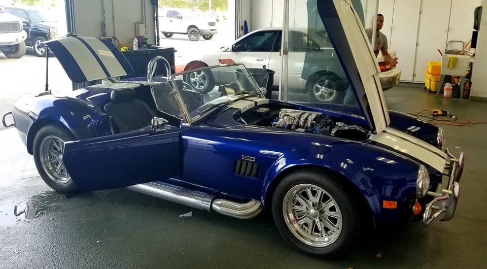 Blue Shelby Cobra roadster with white racing stripes, doors and hood open in garage.