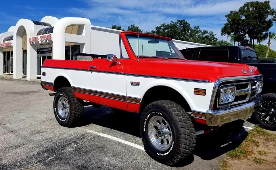Red and white convertible truck with large tires parked in front of a white building on a sunny day.