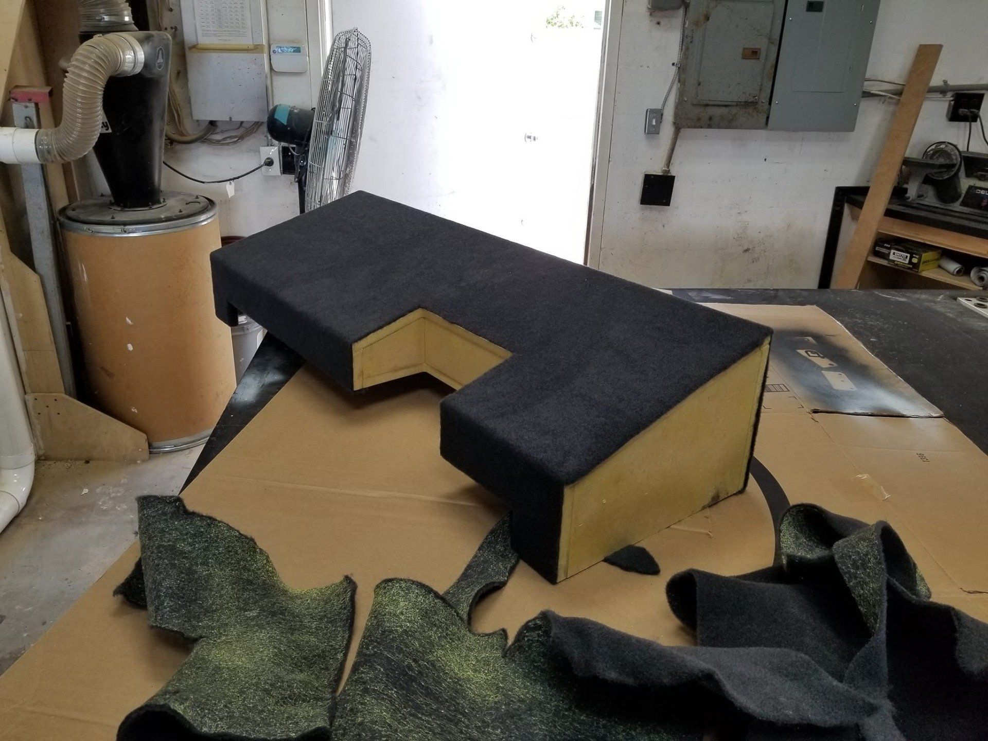 A black carpet-covered L-shaped speaker box with cut pieces of matching carpet on a workbench.