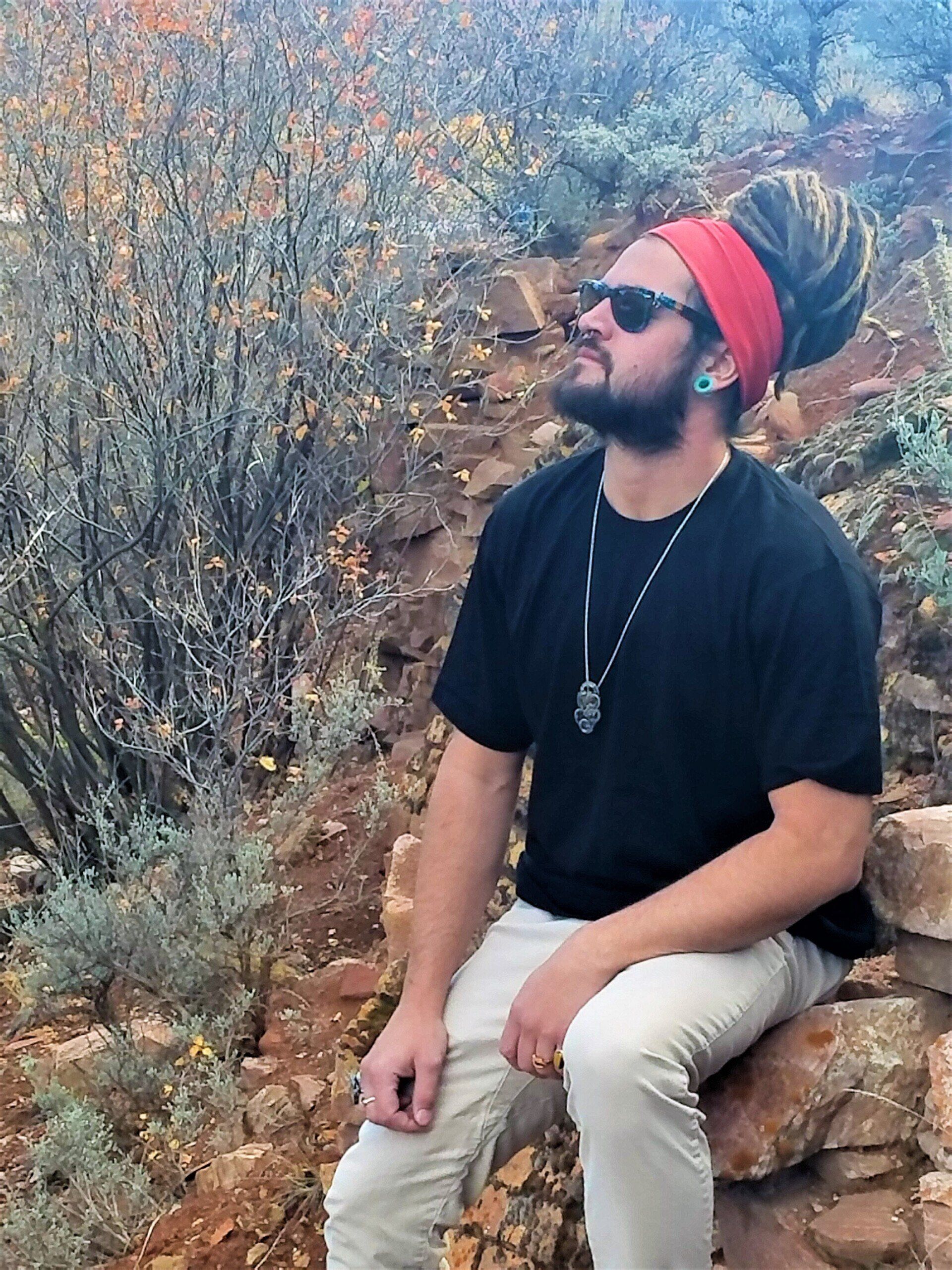 A man with dreadlocks and sunglasses is sitting on a rock