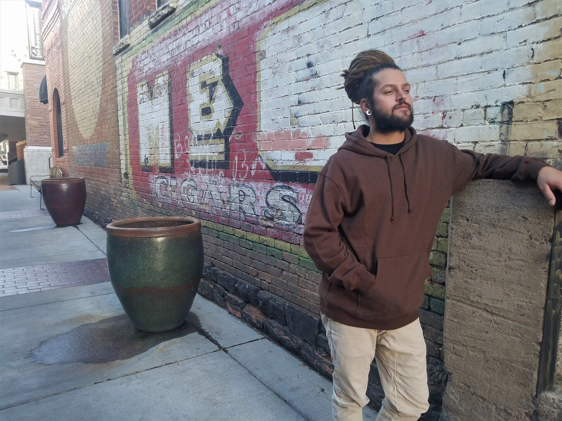 A man in a brown hoodie is leaning against a wall.