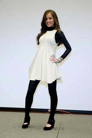 A woman in a white dress and black tights is standing in front of a white wall.