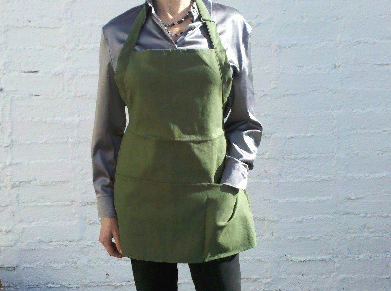 A woman wearing a green apron is standing in front of a white wall