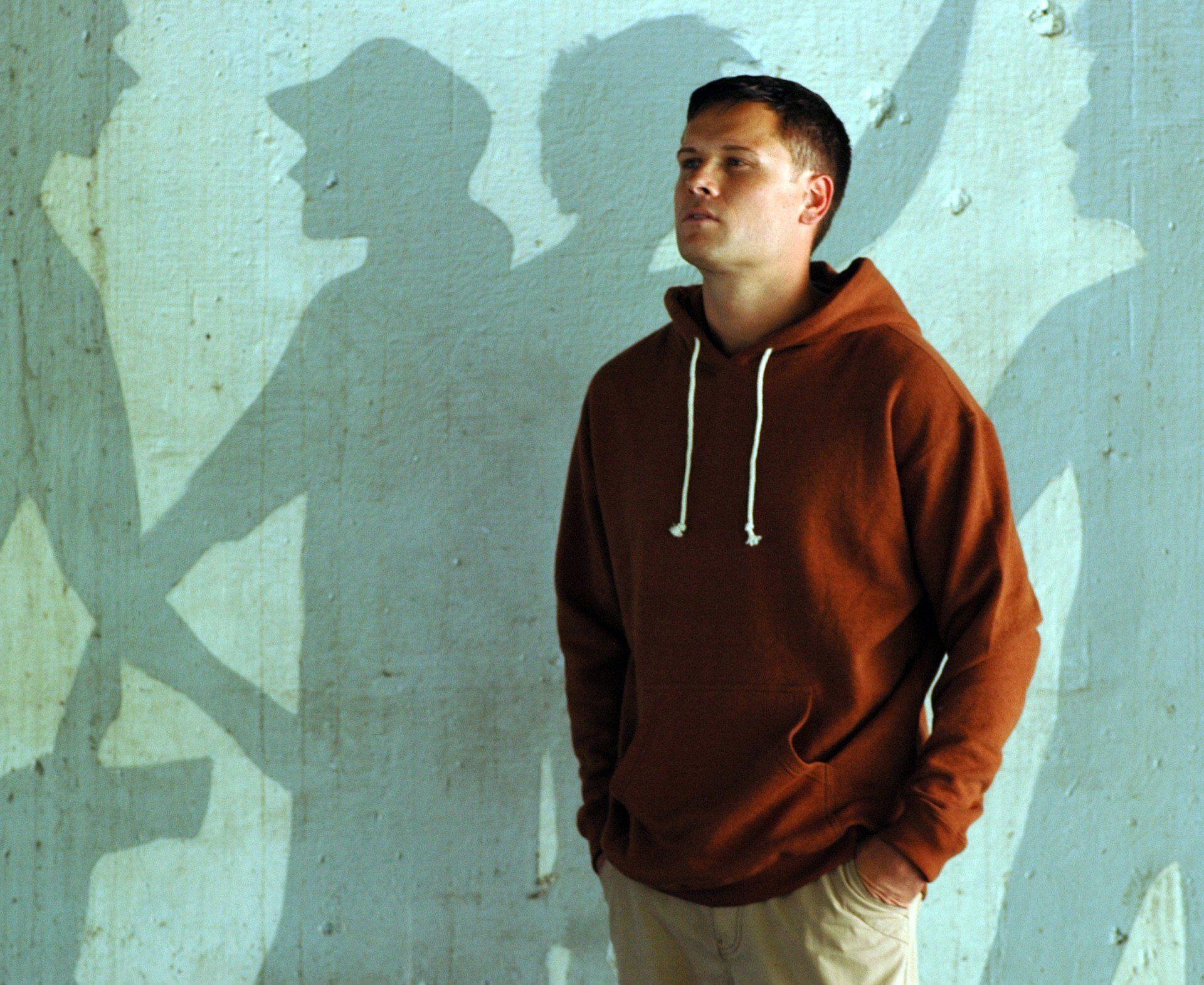 A man in a brown hoodie stands in front of a wall with his hands in his pockets