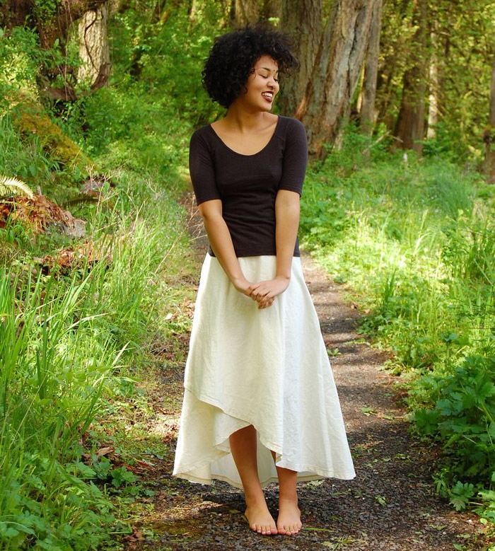 A woman wearing a black shirt and a white skirt is standing on a path in the woods.