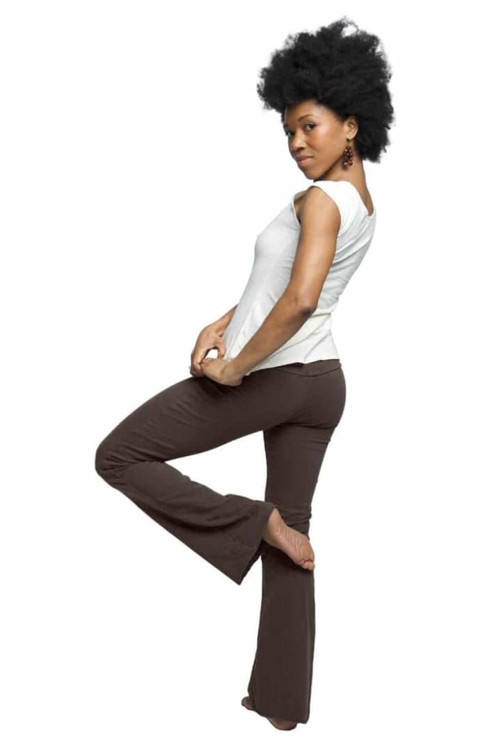 A woman in a white tank top and brown pants is standing on one leg.