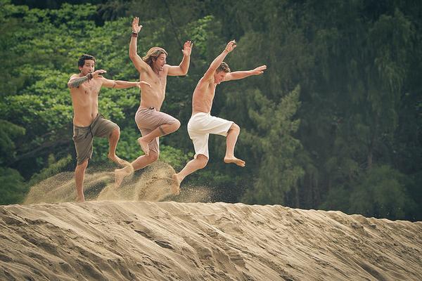 Three men are jumping in the air on top of a sand dune.