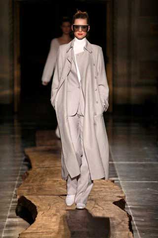 A model is walking down the runway at a fashion show wearing a long coat and sunglasses.