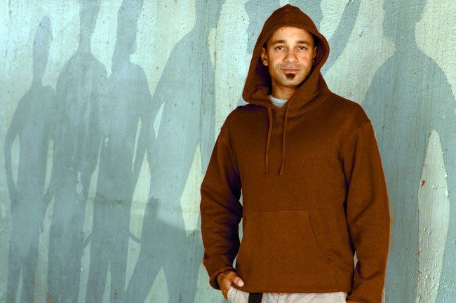 A man in a brown hoodie stands in front of a wall with shadows on it