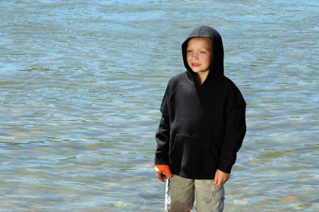 A young boy in a black hoodie is standing in the water.