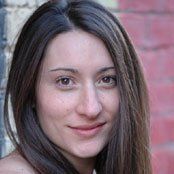 A woman with long hair is smiling in front of a red brick wall.