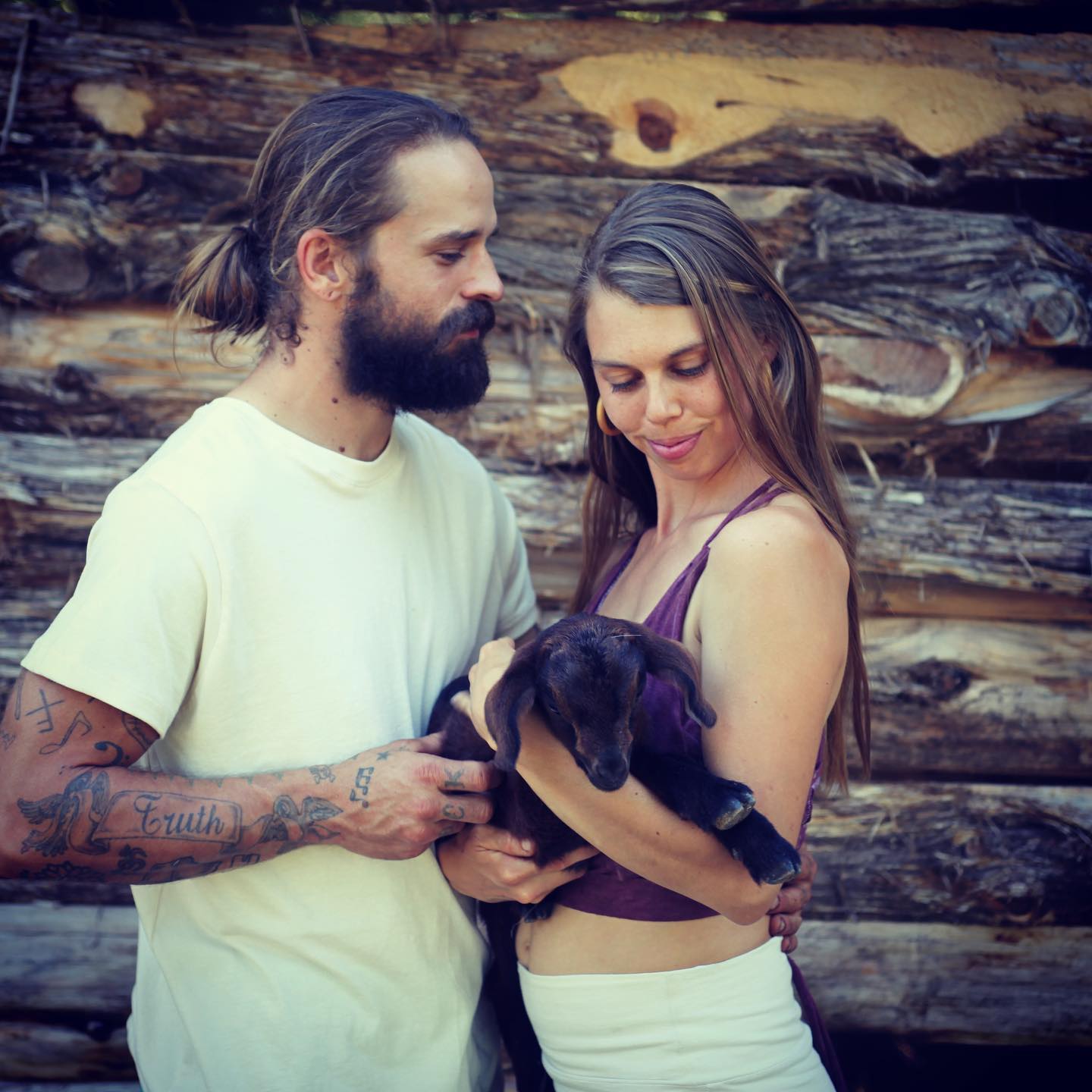 A man with a tattoo on his arm holds a baby goat next to a woman