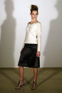 A woman wearing a white top and a black skirt is standing in front of a white wall.