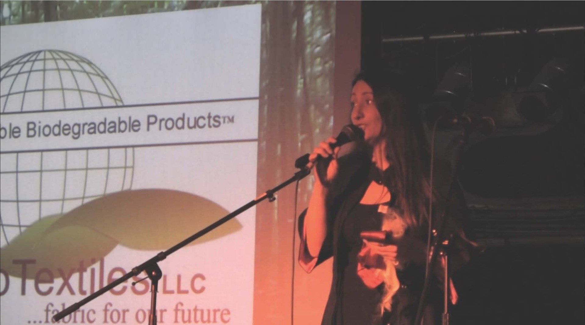 A woman singing into a microphone in front of a sign that says biodegradable products