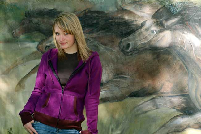 A woman in a purple jacket is standing in front of a painting of horses.