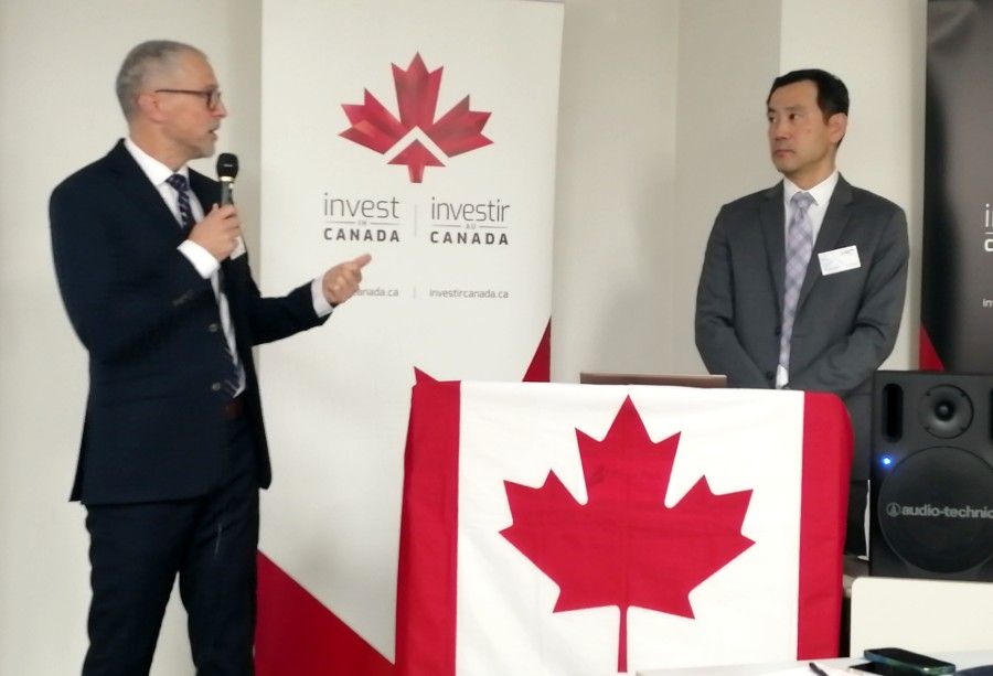 Two men at a podium with a Canadian flag and Invest Canada backdrop. One speaks, pointing, the other listens.
