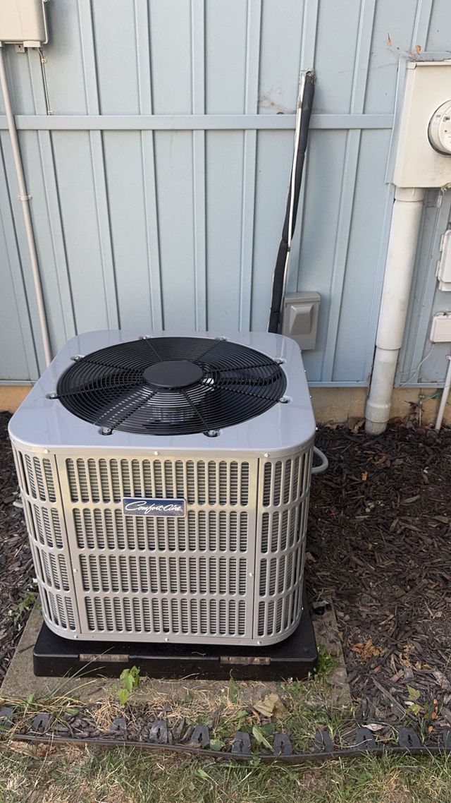 An air conditioning unit outdoors, set on a black platform against a light blue fence.