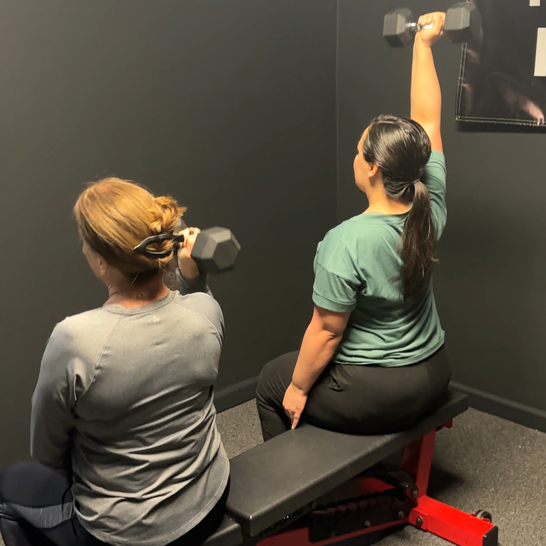 Two women are sitting on a bench lifting dumbbells