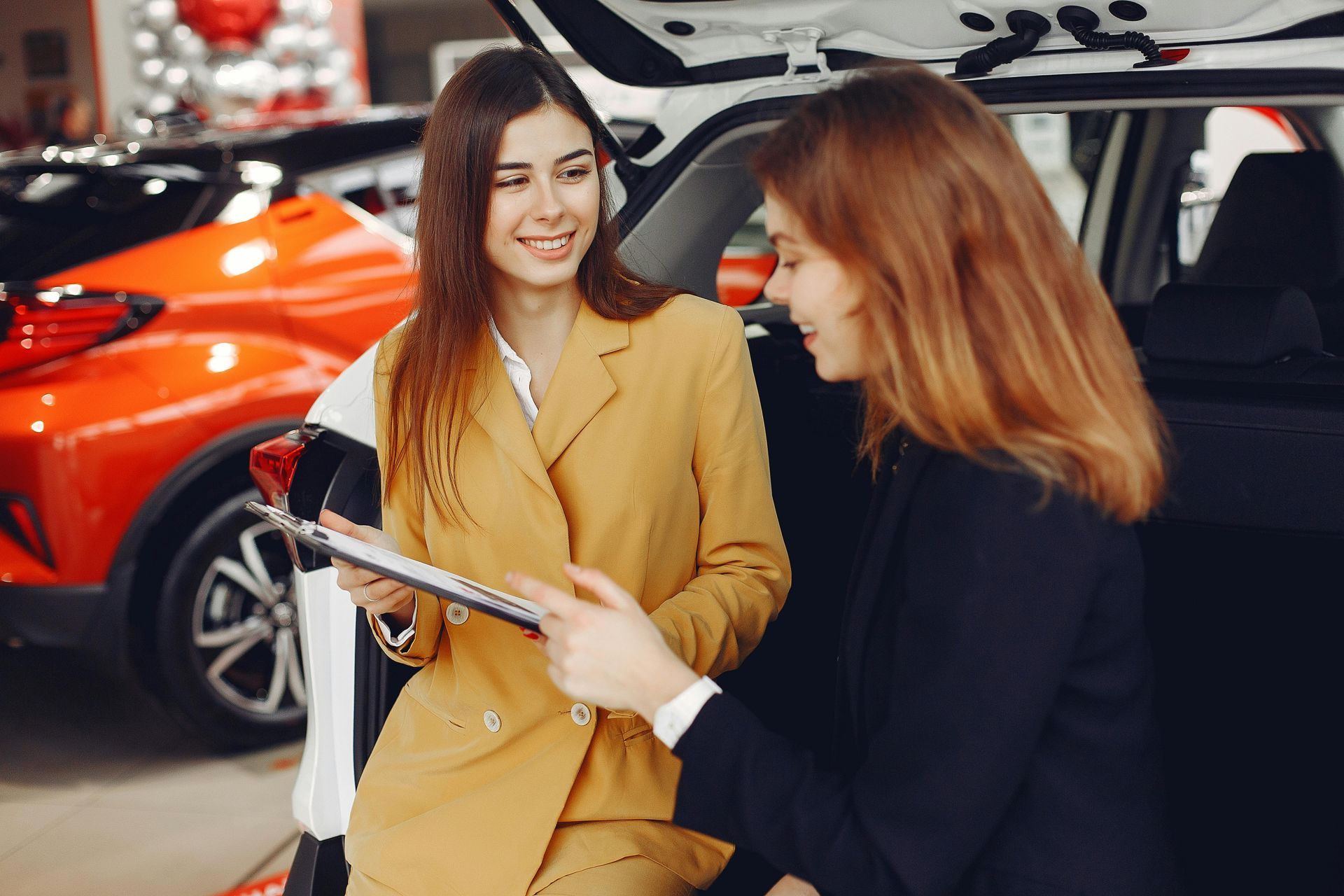 Two women are sitting in the back of a car in a car showroom.