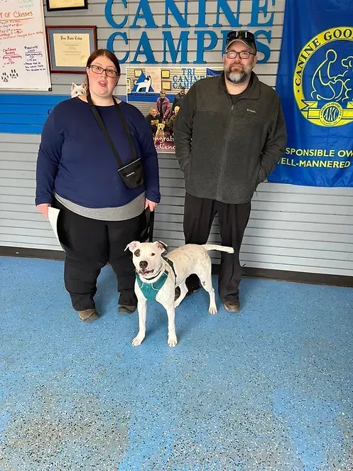 A man and a woman are standing next to a dog in front of a canine campus banner.
