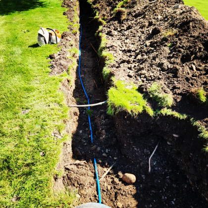 A trench in a grassy lawn with blue pipes running through it. A power saw sits nearby.