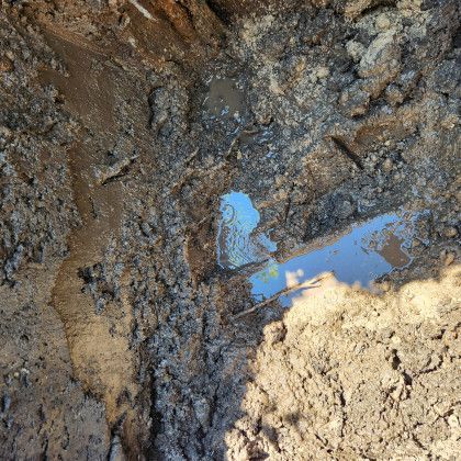 Muddy trench with standing water, possibly rainwater, reflecting blue sky and surrounding earth.