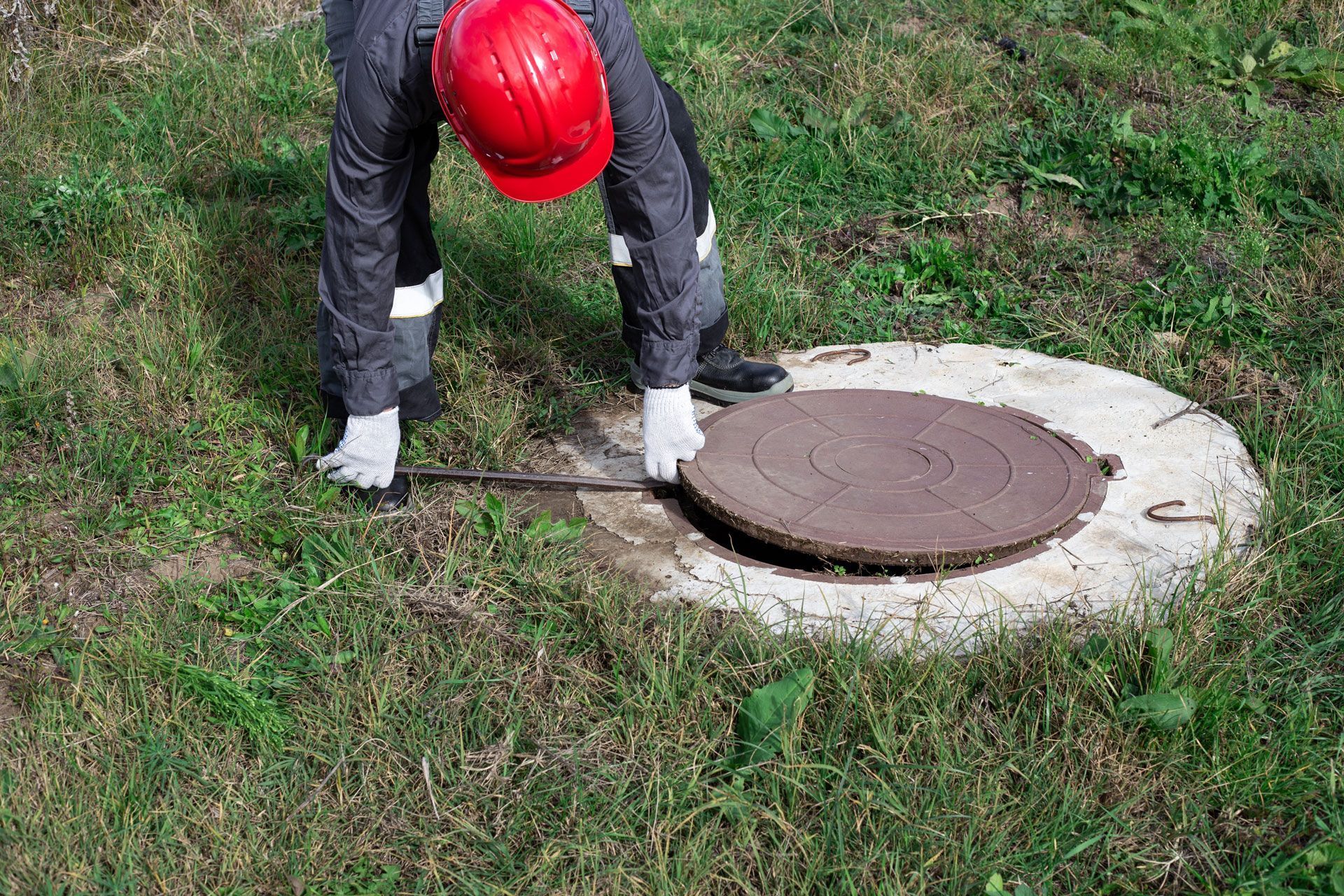 Worker in red hard hat lifting a manhole cover in a grassy area