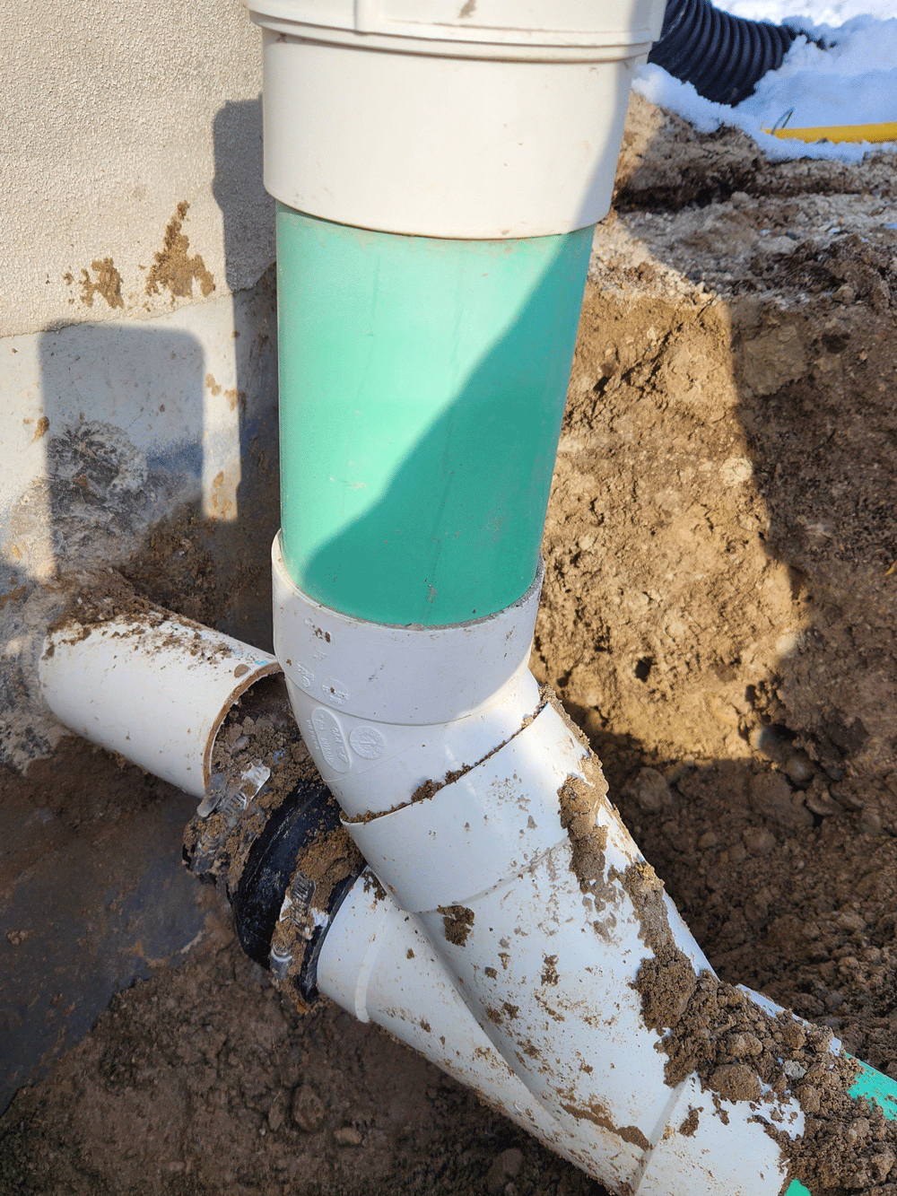 White and green outdoor pipe with broken black coupling in dirt near a house foundation