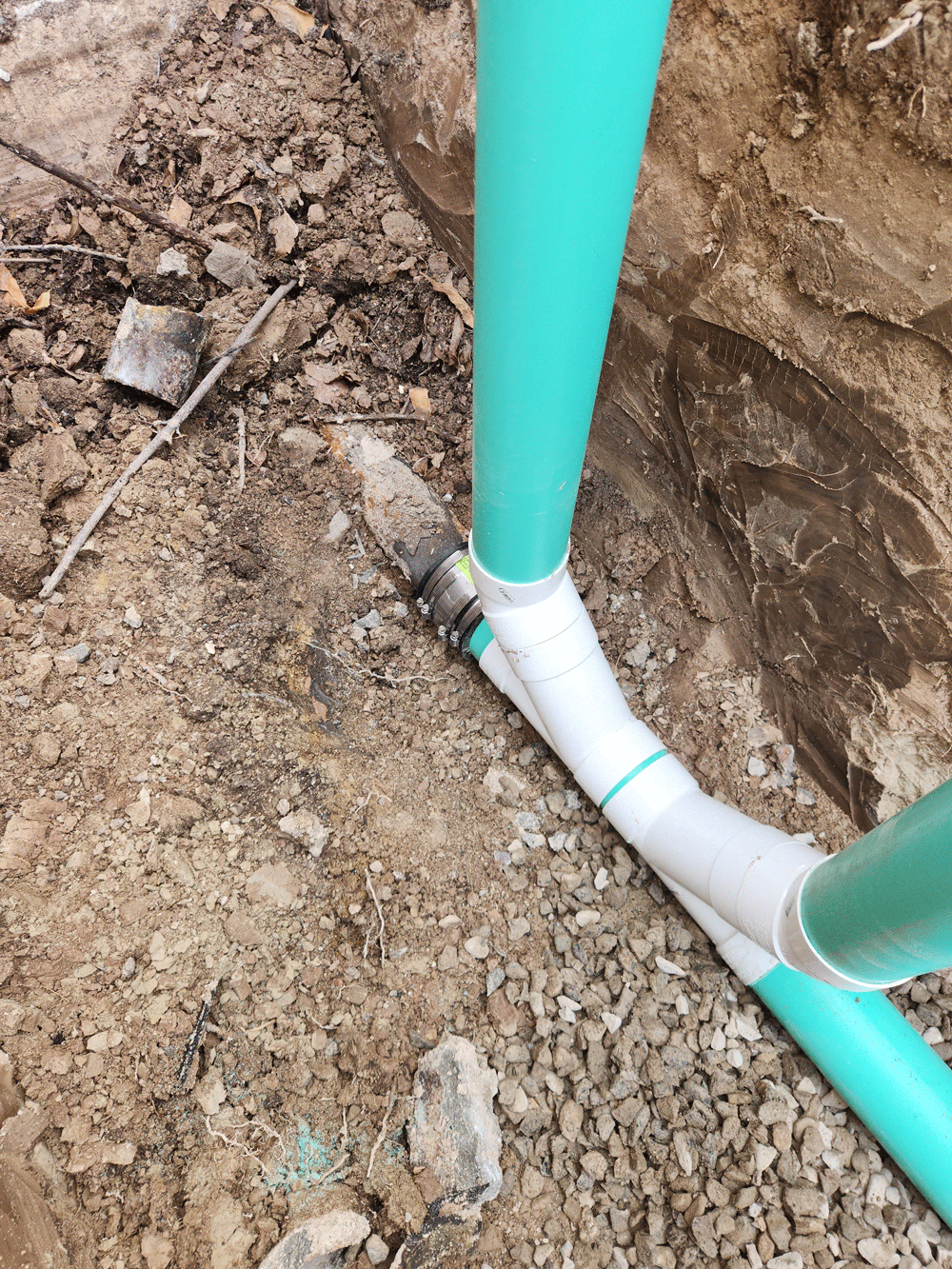 Trench with white PVC pipe connected to turquoise pipes in dirt soil