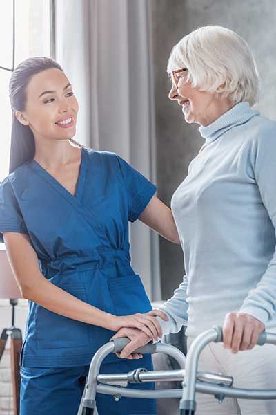 Young female social worker helping senior woman to walk with walker at home 