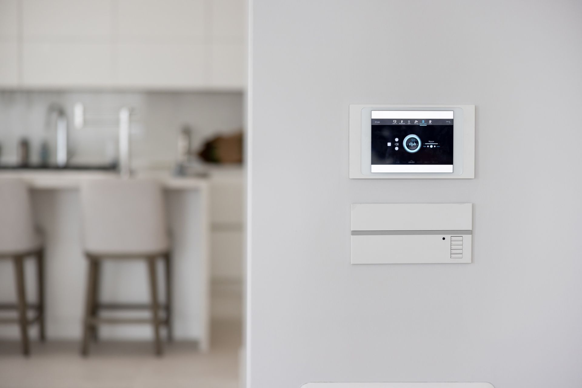 Parete bianca con pannello di controllo per la casa intelligente e citofono in basso, cucina sullo sfondo.