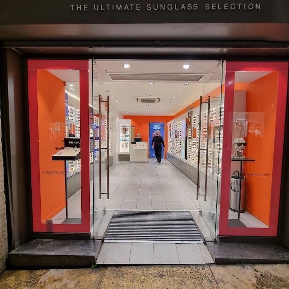 Ingresso di un negozio di occhiali da sole, facciata arancione e in vetro. Una persona entra.