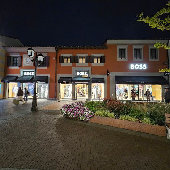 Vista notturna dei negozi BOSS in un centro commerciale all'aperto. Edifici arancioni, vetrine illuminate, gente che cammina.