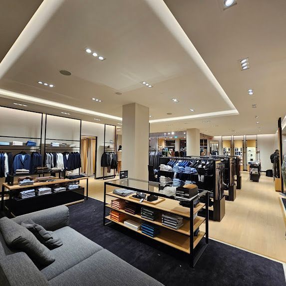 Interno di un negozio di abbigliamento ben illuminato, con scaffali pieni di abiti da uomo. Un divano è posizionato di fronte.