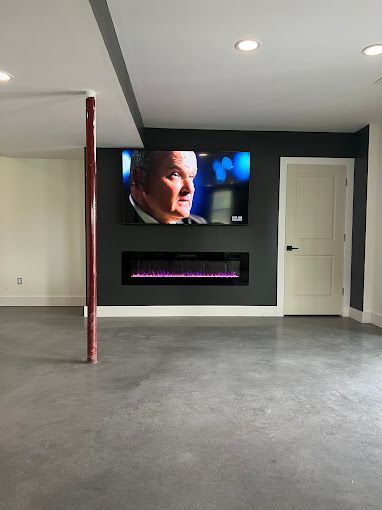 Empty basement room with TV above electric fireplace; gray walls, door on right, exposed red pole on left.