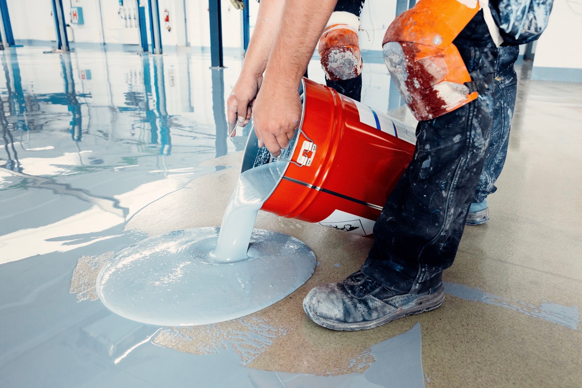 Person pouring light gray epoxy from a red bucket onto a floor during construction.
